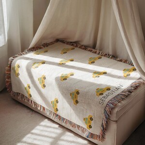 May include: A cream-colored throw blanket featuring a repeating pattern of yellow taxi cabs. The blanket has a fringed edge with multiple colors and the name "SEBASTIAN" is woven into the fabric. The blanket is draped over a white bench.