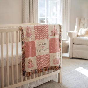 May include: A pink and beige baby blanket with a patchwork design featuring a carriage, castle, crown, and ballerina. The blanket is personalized with the name "Olivia" and the date "06-07-2025". The blanket is draped over a white crib.