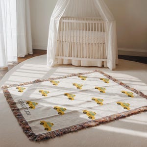 May include: A cream-colored woven baby blanket featuring a repeating pattern of yellow taxi cabs. The blanket has a fringed edge with a mix of colors. The name "SEBASTIAN" is printed in black on the blanket.