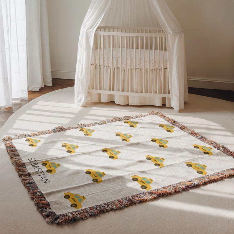 May include: A cream-colored woven baby blanket featuring a repeating pattern of yellow taxi cabs. The blanket has a fringed edge with a mix of colors. The name "SEBASTIAN" is printed in black on the blanket.