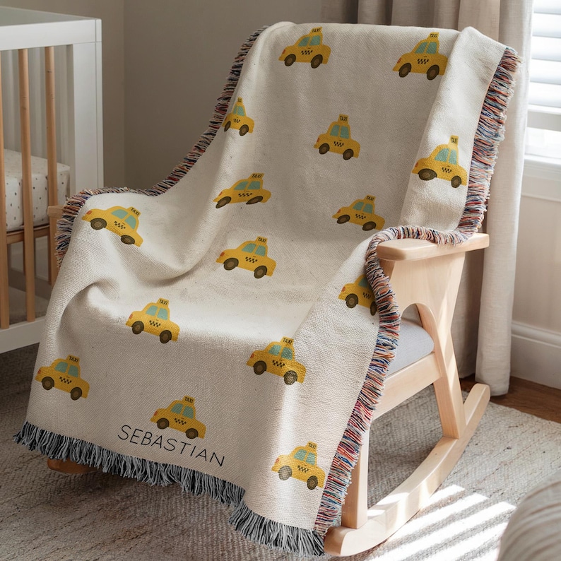 May include: Cream-colored baby blanket with a repeating pattern of yellow taxi cabs. The blanket has a fringed edge with multiple colors and the name "SEBASTIAN" is printed on the bottom.
