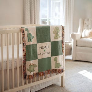 May include: A green and beige dinosaur-themed baby blanket with a fringe. The blanket features a checkerboard pattern with dinosaur illustrations and the name "NOAH" with a heart and the date "07-06-2025". The blanket is draped over a white cot.
