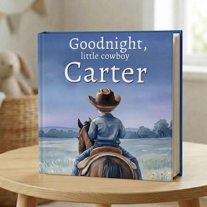 Pode incluir: Um livro infantil com o t&iacute;tulo "Goodnight, little cowboy Carter" na capa. A ilustra&ccedil;&atilde;o mostra um jovem cowboy montado a cavalo contra um c&eacute;u azul. O livro est&aacute; numa mesa de madeira. O livro tem uma capa azul.