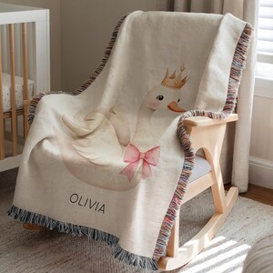 May include: Cream-colored woven baby blanket with a swan design. The swan wears a gold crown and a pink bow. The blanket has a fringe border with multiple colors and the name "OLIVIA" is printed on the blanket.