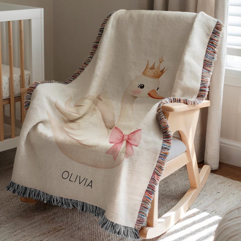 May include: Cream-colored woven baby blanket with a swan design. The swan wears a gold crown and a pink bow. The blanket has a fringe border with multiple colors and the name "OLIVIA" is printed on the blanket.