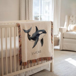 Puede incluir: Una manta de bebé beige con una ilustración de acuarela de una orca en blanco y negro. La manta tiene un borde con flecos con hilos rojos, naranjas y marrones. El nombre "LUCAS" está impreso en letras negras debajo de la ballena.