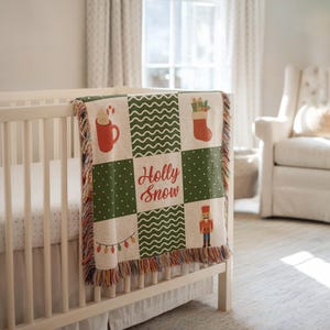 May include: A Christmas-themed baby blanket draped over a crib. The blanket features a patchwork design with festive images like a mug, stocking, and nutcracker. The words "Holly Snow" are written in red. The blanket has a fringed edge.