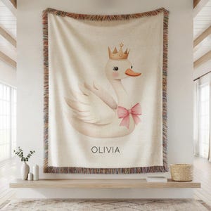 May include: Cream-colored throw blanket featuring a swan illustration wearing a gold crown and a pink bow. The name "OLIVIA" is printed below the swan. The blanket has a fringed border.
