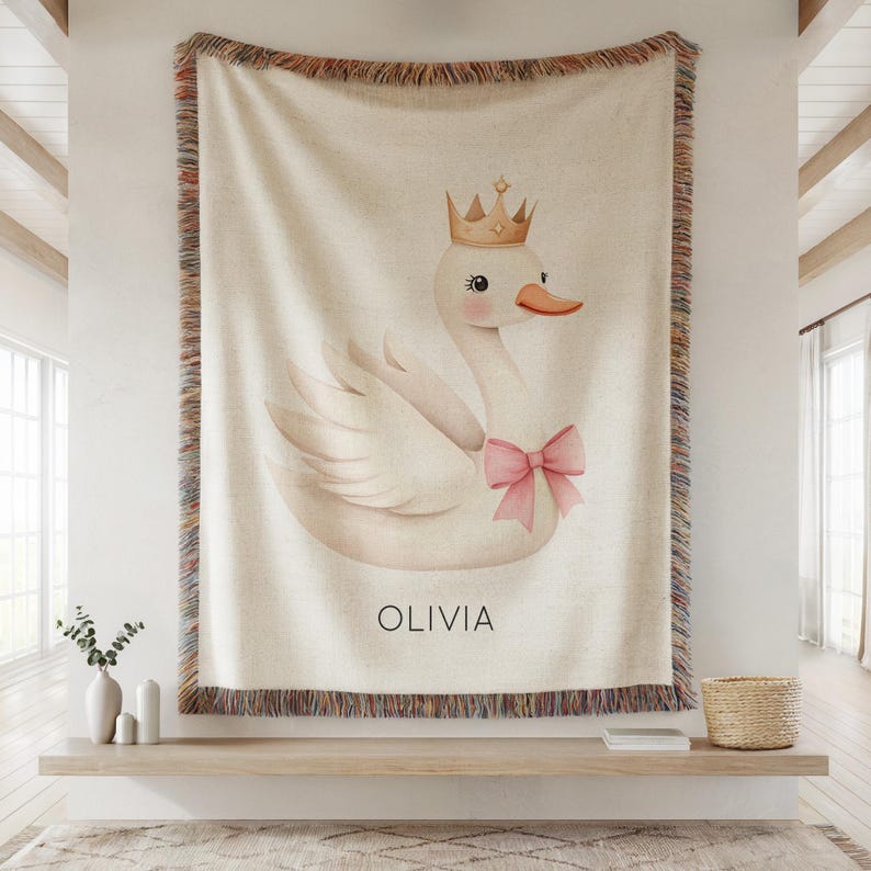 May include: Cream-colored throw blanket featuring a swan illustration wearing a gold crown and a pink bow. The name "OLIVIA" is printed below the swan. The blanket has a fringed border.