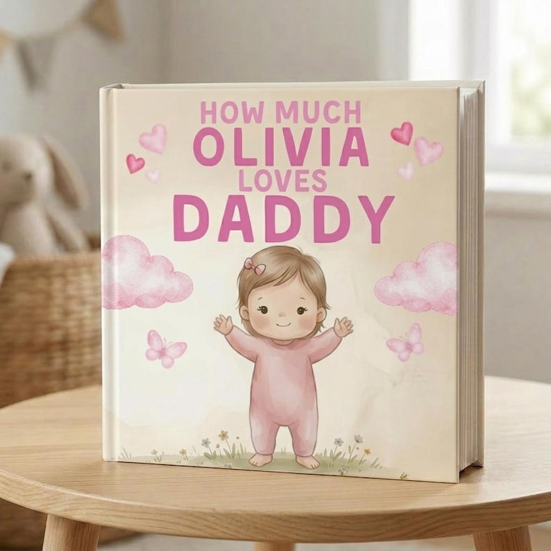 Peut inclure: Livre personnalis&eacute; pour enfants avec le titre "How Much Olivia Loves Daddy" en rose. La couverture pr&eacute;sente une illustration de b&eacute;b&eacute; en barboteuse rose, entour&eacute;e de c&oelig;urs, de nuages et de papillons roses. Le livre est pos&eacute; sur une table en bois.