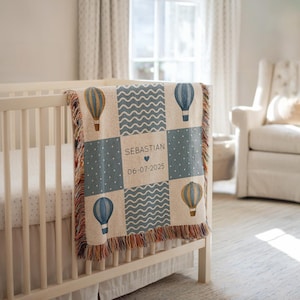 以下が含まれることがあります： 青、ベージュ、白の熱気球、波線、水玉模様が特徴のパッチワークデザインの織りベビーブランケット。「SEBASTIAN」という名前と「06-07-2025」の日付でパーソナライズされています。ブランケットはフリンジエッジです。