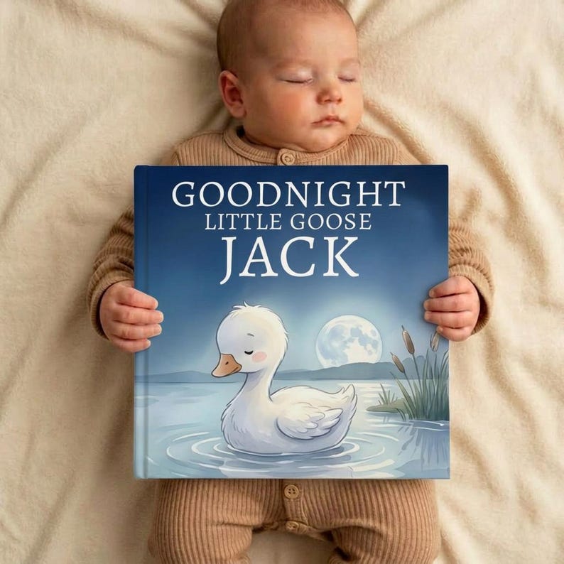 May include: A baby lies peacefully, holding a children's book. The book cover features a serene illustration of a white goose on a lake under a full moon. The title reads "GOODNIGHT LITTLE GOOSE JACK". The baby is wearing a brown onesie.