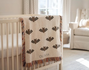 May include: A cream-colored baby blanket with a repeating pattern of brown bats. The blanket is draped over a white crib and has a fringed edge with multiple colors. The name "OLIVIA" is printed on the blanket.