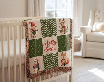 May include: A Christmas-themed woven baby blanket on a crib. The blanket has squares with festive illustrations of an elf, reindeer, penguin, and fox. The words "Holly Snow" are in the center. The blanket has a fringed edge.