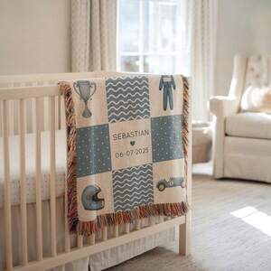 Könnte beinhalten: Eine gewebte Babydecke mit Rennsport-Motiv, über einem Kinderbett drapiert. Die Decke zeigt Quadrate mit Bildern zum Thema Rennsport, darunter eine Trophäe, ein Rennwagen und ein Helm. Der Name "Sebastian" und ein Datum sind ebenfalls abgebildet.