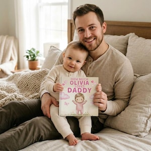 Peut inclure: Un livre pour enfants personnalis&eacute; intitul&eacute; &laquo;&nbsp;How Much Olivia loves Daddy&nbsp;&raquo;. La couverture pr&eacute;sente une illustration de b&eacute;b&eacute; en rose, avec des nuages et des c&oelig;urs roses. Le livre est tenu par un b&eacute;b&eacute; et un homme.