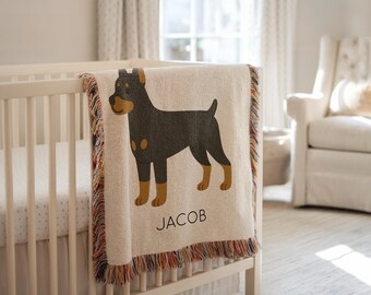 Puede incluir: Una manta para bebé de color crema con un diseño tejido que presenta una ilustración de un perro negro y marrón. La manta está sobre una cuna blanca y tiene un borde con flecos con hilos coloridos. El nombre "JACOB" está impreso en negro.