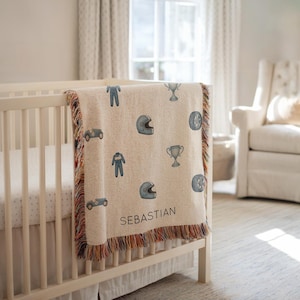 Könnte beinhalten: Eine beige gewebte Babydecke mit Rennwagen-Motiv. Die Decke zeigt blaue Rennwagen, Helme, Rennanzüge und Trophäen. Der Name "Sebastian" ist in Schwarz aufgedruckt. Die Decke hat einen Fransenrand.