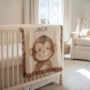 Op de afbeelding: Een beige babydeken met een cartoon aap illustratie en de naam "JACK" in zwarte letters. De deken heeft een gefranjerde rand met meerdere kleuren en is over een witte wieg gedrapeerd.