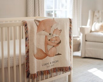 May include: A cream-colored woven baby blanket featuring a watercolor illustration of a mother fox embracing a baby fox. The blanket has a fringed edge with multiple colors and the text "LIAM + MOM EST. 2025". The blanket is draped over a white crib.