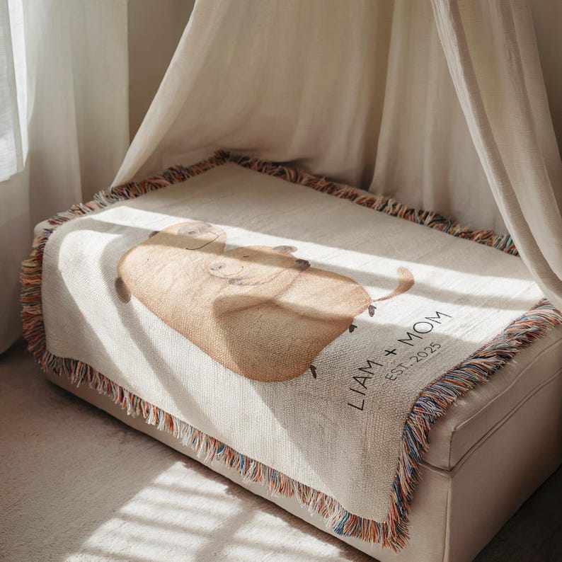 May include: A beige woven throw blanket with a cartoon illustration of two pigs. The blanket has a fringed edge with multiple colors. The text "LIAM + MOM EST. 2025" is printed on the blanket.