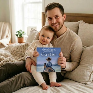 Pode incluir: Um pai e um beb&ecirc; est&atilde;o sentados em uma cama, ambos sorrindo para a c&acirc;mera. O beb&ecirc; segura um livro intitulado "Goodnight, little cowboy Carter" com uma ilustra&ccedil;&atilde;o de um cowboy a cavalo. O livro tem uma capa azul. O pai usa uma camisa bege de manga comprida.