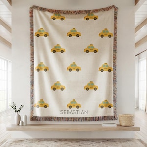 May include: Cream-colored throw blanket featuring a repeating pattern of yellow taxi cabs. The blanket has a fringed edge with a mix of colors. The name "SEBASTIAN" is printed in black at the bottom of the blanket.