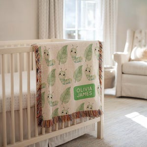 May include: A cream-colored baby blanket with a repeating pattern of green caterpillars and leaves. The blanket has a fringed edge with multiple colors. The name "OLIVIA JAMES" is printed on a green rectangle.