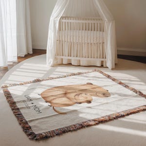May include: A beige woven throw blanket with a watercolor illustration of two capybaras. The blanket has a fringed edge with multiple colors. Text on the blanket reads "LIAM + MOM EST. 2025". A white crib with a canopy is in the background.