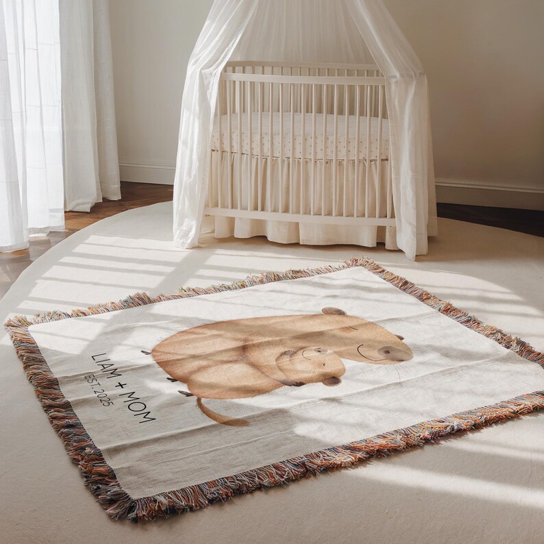 May include: A beige woven throw blanket with a watercolor illustration of two capybaras. The blanket has a fringed edge with multiple colors. Text on the blanket reads "LIAM + MOM EST. 2025". A white crib with a canopy is in the background.