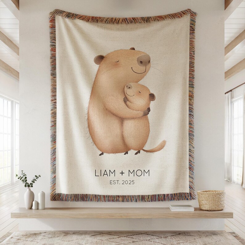 May include: A beige throw blanket featuring a heartwarming illustration of two cartoon animals embracing. The blanket has a fringed border with brown, orange, and beige hues. Text on the blanket reads "LIAM + MOM EST. 2025".
