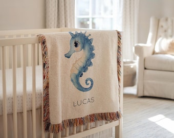 May include: A cream-colored baby blanket with a blue seahorse design and the name "LUCAS" in black. The blanket has a fringed edge with red, orange, and brown threads. It is draped over a white crib.