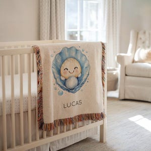 May include: A cream-colored baby blanket with a colorful fringe, draped over a crib. The blanket features a watercolor illustration of a baby in a blue seashell, with the name "LUCAS" printed below.