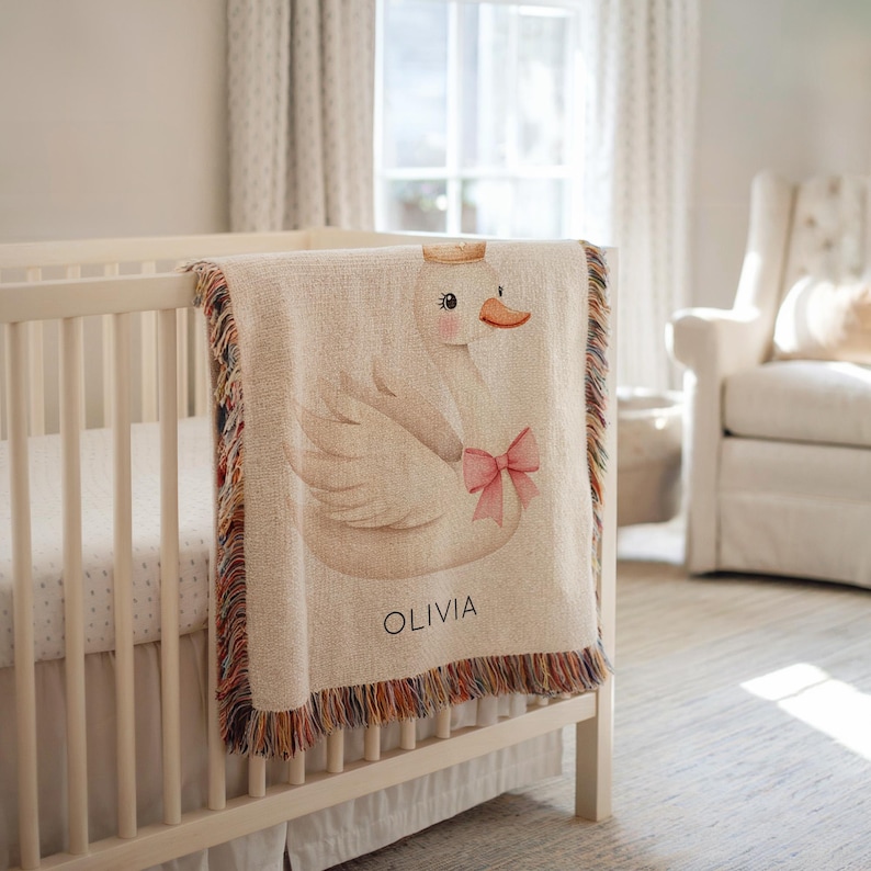 May include: A beige baby blanket with a swan design, a pink bow, and the name "OLIVIA" printed in black. The blanket has a fringed edge with multiple colors and is draped over a white crib.
