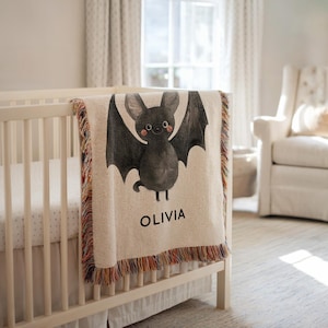 Puede incluir: Una manta para bebé de color crema con una ilustración de un murciélago. El murciélago es gris con alas grandes y una cara sonriente. El nombre "OLIVIA" está impreso en negro debajo. La manta tiene un borde con flecos con hilos coloridos.