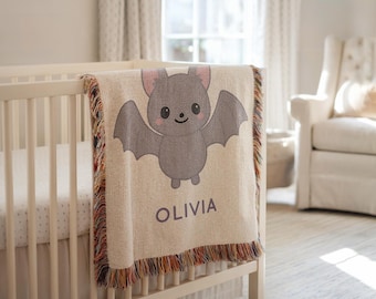 May include: A beige baby blanket with a cartoon bat design and the name "OLIVIA" in purple lettering. The blanket has a fringed edge with multiple colors and is draped over a white crib.
