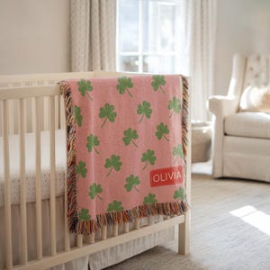 May include: A pink baby blanket with a green shamrock pattern draped over a white crib. The blanket has a fringed edge and a red rectangle with the name "OLIVIA" printed on it. The blanket is made of a soft material.