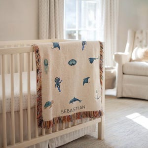 May include: A beige baby blanket with a colourful fringe is draped over a cot. The blanket features blue watercolour illustrations of sea creatures, including a diver, stingrays, and a dolphin. The name "SEBASTIAN" is printed on the blanket.
