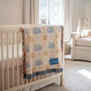 May include: A cream-colored baby blanket with a repeating pattern of blue clouds, yellow suns, and small stars. The blanket has a blue fringe and a blue patch with the name "LUKE" embroidered on it. The blanket is draped over a white crib.