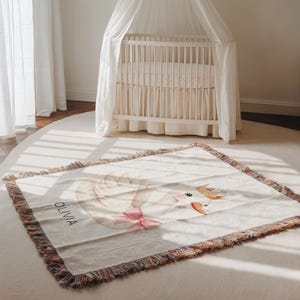 May include: A cream-colored baby blanket with a swan design and the name "OLIVIA" printed on it. The swan has a pink bow and a gold crown. The blanket has a fringed edge and is placed on a white rug.