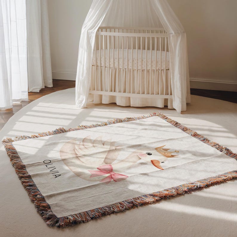 May include: A cream-colored baby blanket with a swan design and the name "OLIVIA" printed on it. The swan has a pink bow and a gold crown. The blanket has a fringed edge and is placed on a white rug.