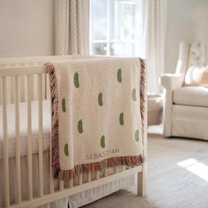 May include: A cream-colored baby blanket with a repeating pattern of green pickle illustrations. The blanket has a fringed edge with multiple colors and the name "SEBASTIAN" is printed on the blanket.