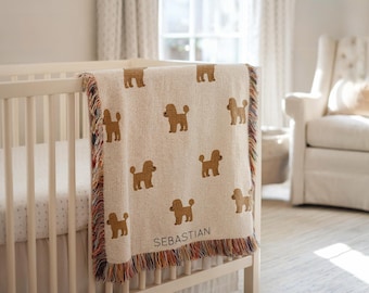 May include: A cream-colored baby blanket with brown poodle designs and the name "Sebastian" woven into the fabric. The blanket has a fringed edge with multicolored threads and is draped over a white crib.
