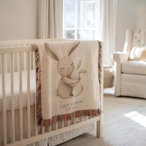 Puede incluir: Una manta de bebé beige con una ilustración en acuarela de dos conejos abrazándose. La manta tiene un borde con flecos multicolores. El texto en la manta dice "LIAM + MOM EST. 2025". La manta está sobre una cuna blanca.