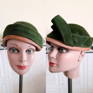 Op de afbeelding: Twee groene vilt hoeden met bruine bies. Een hoed heeft een grote groene strik bovenop. Beide hoeden zijn op mannequins met neutrale make-up.