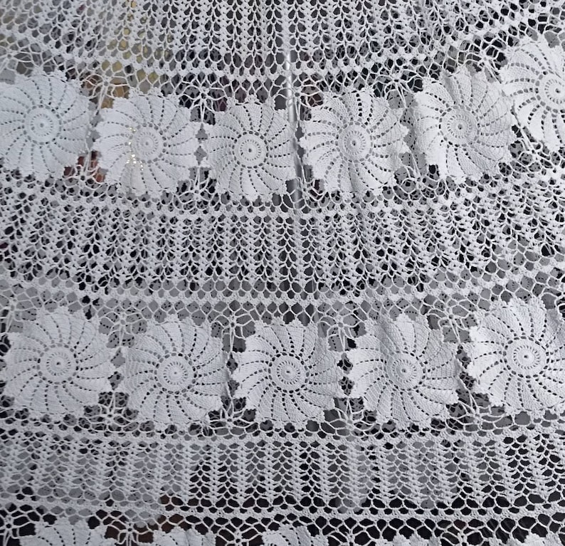 May include: White crocheted tablecloth with a delicate floral pattern. The tablecloth features a repeating design of small, intricate flowers.