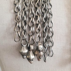 May include: Close-up of a silver-toned necklace with multiple strands of linked chain. The chain has a unique looped design, and each strand ends with a small, decorative pendant. The pendants have a bulbous shape.