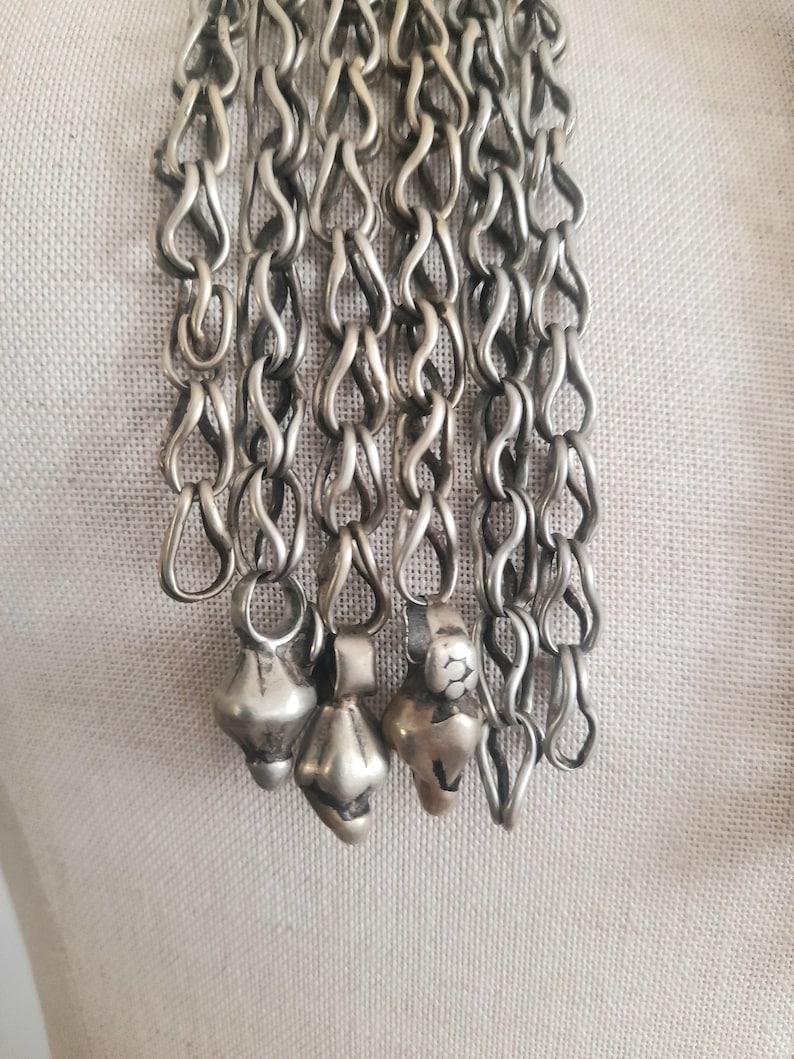 May include: Close-up of a silver-toned necklace with multiple strands of linked chain. The chain has a unique looped design, and each strand ends with a small, decorative pendant. The pendants have a bulbous shape.