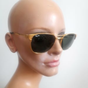May include: Gold-framed sunglasses with dark lenses. The Ray-Ban logo is visible on the upper left lens. The sunglasses are displayed on a mannequin head.