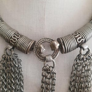May include: Silver-toned necklace with intricate detailing. The necklace features a coiled design on the upper portion, connected to a central circular element. Dangling from the necklace are multiple chains, creating a layered effect. The piece is displayed on a mannequin.